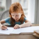 A young girl with red hair focused on drawing at a wooden desk indoors.