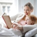 Cheerful young woman hugging cute little girl and reading book together while lying in soft bed in light bedroom at home in daytime