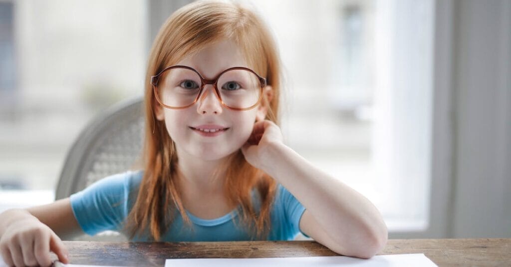 Smiling girl wearing eyeglasses, sitting indoors and doing homework at a table.