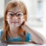 Smiling girl wearing eyeglasses, sitting indoors and doing homework at a table.