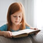 A young girl with red hair is engrossed in reading a book indoors, creating a serene and studious atmosphere.