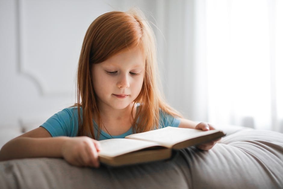 A young girl with red hair is engrossed in reading a book indoors, creating a serene and studious atmosphere.