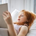 A young girl relaxing in bed by the window, enjoying a book and embracing leisure time.