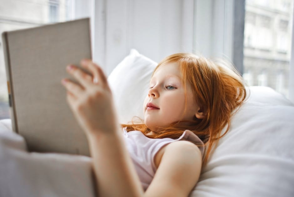 A young girl relaxing in bed by the window, enjoying a book and embracing leisure time.