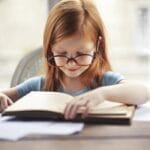 A young girl with glasses intently reads a book indoors, embodying the essence of learning and discovery.