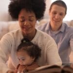 A family of three joyfully reading a book together in a warm, comfortable living room setting.