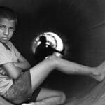 A contemplative young boy sits barefoot in a large industrial pipe in black and white.