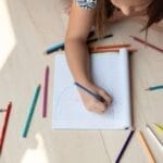 A young child drawing on paper surrounded by colorful pencils, illustrating creativity.