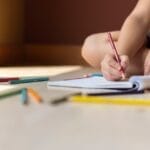 A child sitting indoors, drawing in a notebook with colorful crayons, surrounded by art supplies.