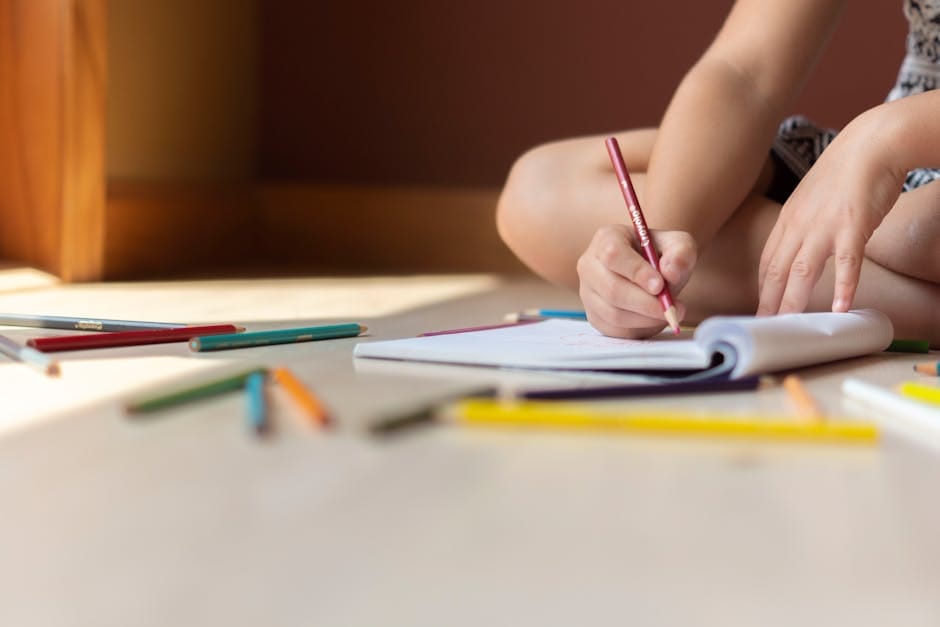 A child sitting indoors, drawing in a notebook with colorful crayons, surrounded by art supplies.