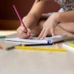 A child seated on the floor using colored pencils to draw on paper, showcasing creativity.