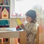 A young girl in a bright classroom engaged in a creative arts and crafts project at a table.