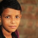 A thoughtful young boy gazes into the camera with a serious expression against a blurred brick background.