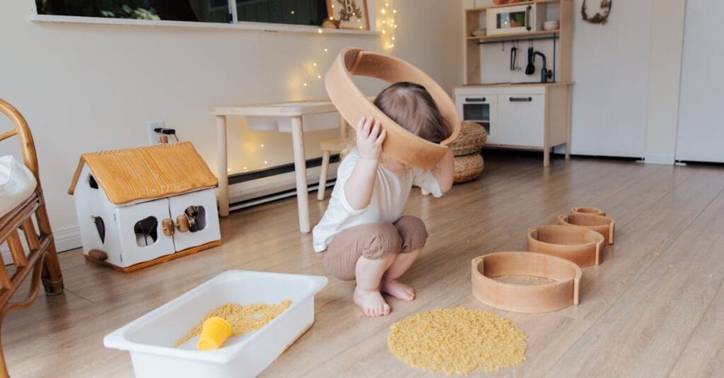 Full length of anonymous toddler squatting barefoot on floor playing with round wooden shapes of different size and pasta and putting biggest shape on while developing fine motor skills at home