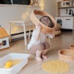 Full length of anonymous toddler squatting barefoot on floor playing with round wooden shapes of different size and pasta and putting biggest shape on while developing fine motor skills at home