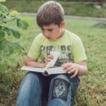 A young boy sits on grass outdoors, engrossed in reading a book.