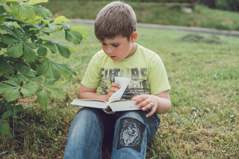 A young boy sits on grass outdoors, engrossed in reading a book.