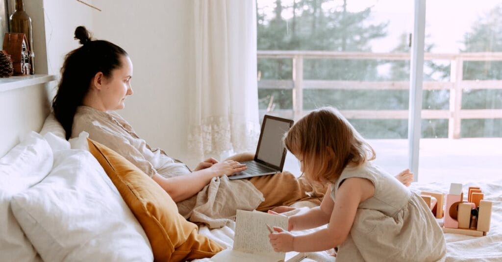 A mother works remotely on a laptop while her child plays next to her in a cozy bedroom setting.