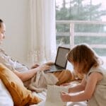 A mother works remotely on a laptop while her child plays next to her in a cozy bedroom setting.