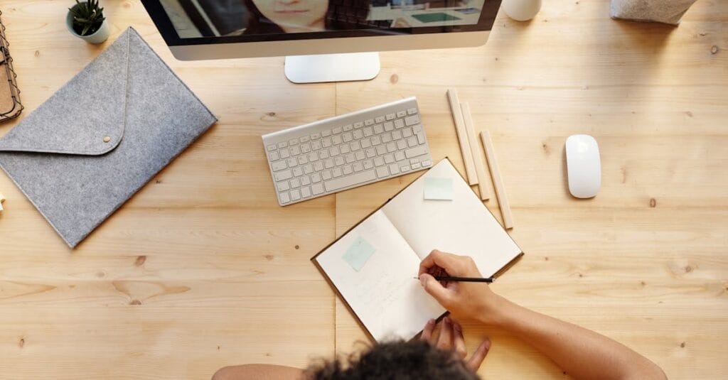 A teenager studying at home using a computer for online learning, taking notes in a notebook.