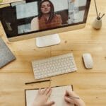 Person taking notes during a video call at a neatly organized home desk, showing remote work lifestyle.