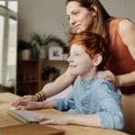 A mother and her child smiling while using a computer at home, focused on learning.