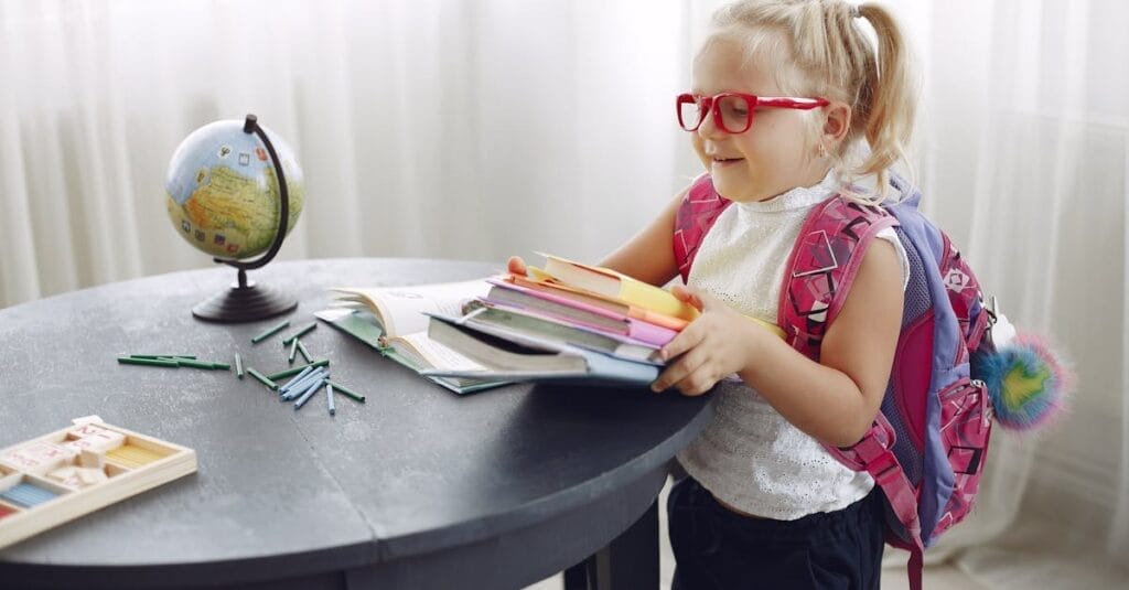 Little girl wearing casual clothes and glasses smiling while preparing books for homework at home during weekend and looking away