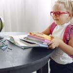 Little girl wearing casual clothes and glasses smiling while preparing books for homework at home during weekend and looking away