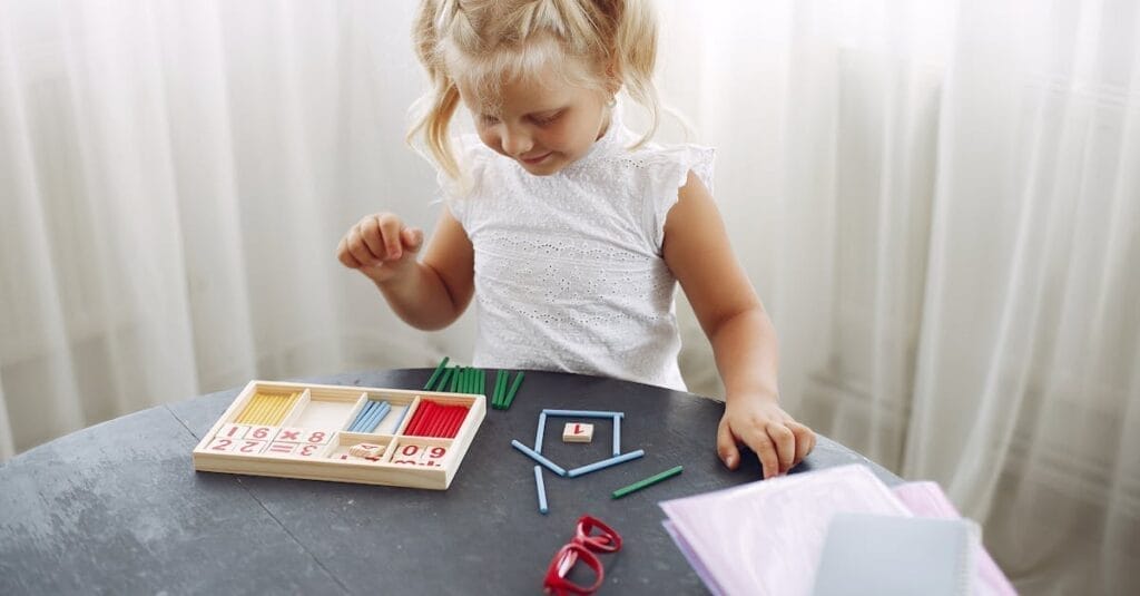 Blond little girl standing while playing with puzzle in living room at home and counting sticks different colors during weekend