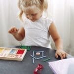 Blond little girl standing while playing with puzzle in living room at home and counting sticks different colors during weekend