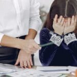 A young girl overwhelmed by homework with a teacher assisting her indoors.