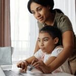 A mother helps her daughter with an online lesson on a laptop, showcasing family bonding and education at home.