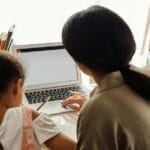 A mother helps her daughter with online learning on a laptop at home, emphasizing family education.