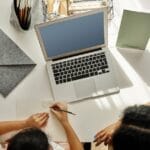 Mother helping daughter with homework at a desk with a laptop indoors.