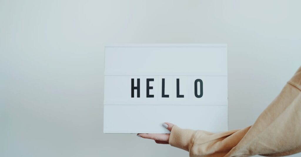 A hand holds a white sign displaying the word 'Hello' against a plain background.