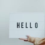 A hand holds a white sign displaying the word 'Hello' against a plain background.