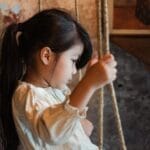 Thoughtful young girl sitting on a swing indoors, in a rustic setting with soft lighting.