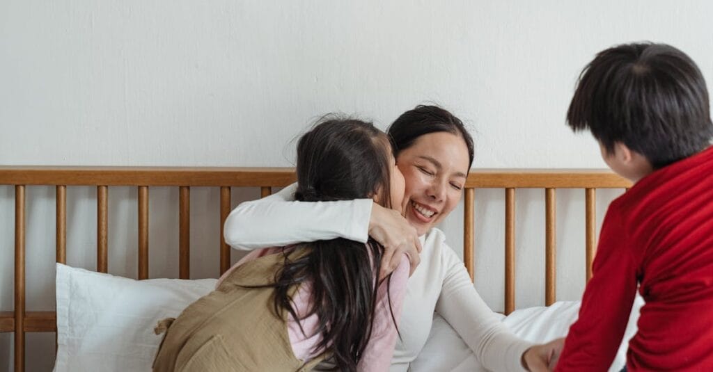 Happy mother in casual clothes sitting on bed hugging little daughter while girl kissing smiling woman on cheek near brother