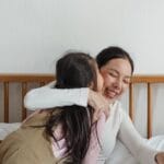 Happy mother in casual clothes sitting on bed hugging little daughter while girl kissing smiling woman on cheek near brother