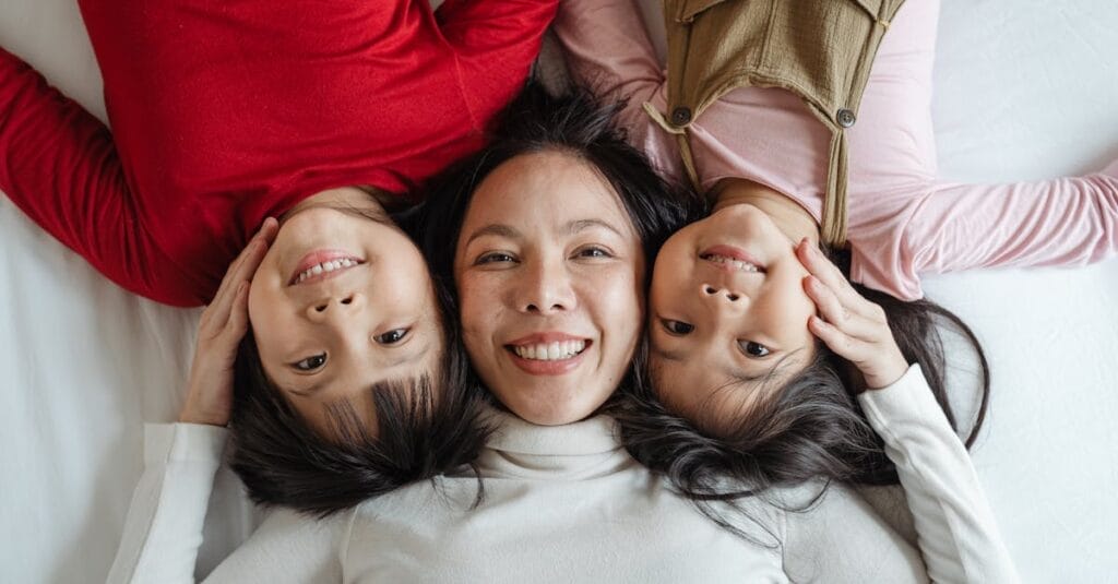 Top view of excited smiling Asian mother caressing adorable little kids in bright casual clothes while resting together on comfortable bed with white sheets and looking at camera