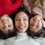 Top view of excited smiling Asian mother caressing adorable little kids in bright casual clothes while resting together on comfortable bed with white sheets and looking at camera