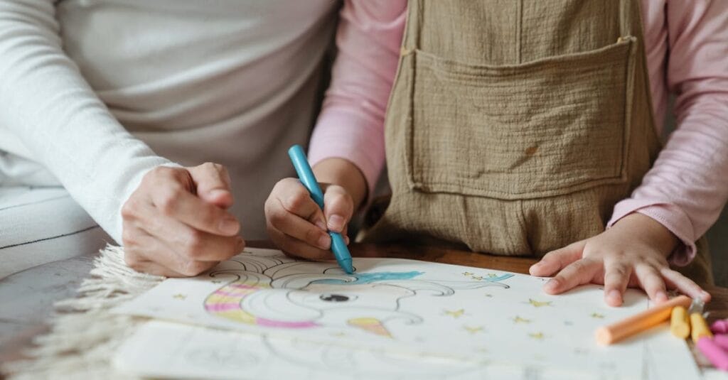 Crop anonymous mother with little child in casual clothes sitting at table and drawing with crayons on papers in daylight