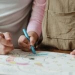 Crop anonymous mother with little child in casual clothes sitting at table and drawing with crayons on papers in daylight