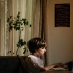 A young boy intensely focused on coding on a computer in a cozy, dimly-lit room.