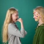 Blonde siblings having fun with colorful candy face stickers on a green background. Artistic play indoors.