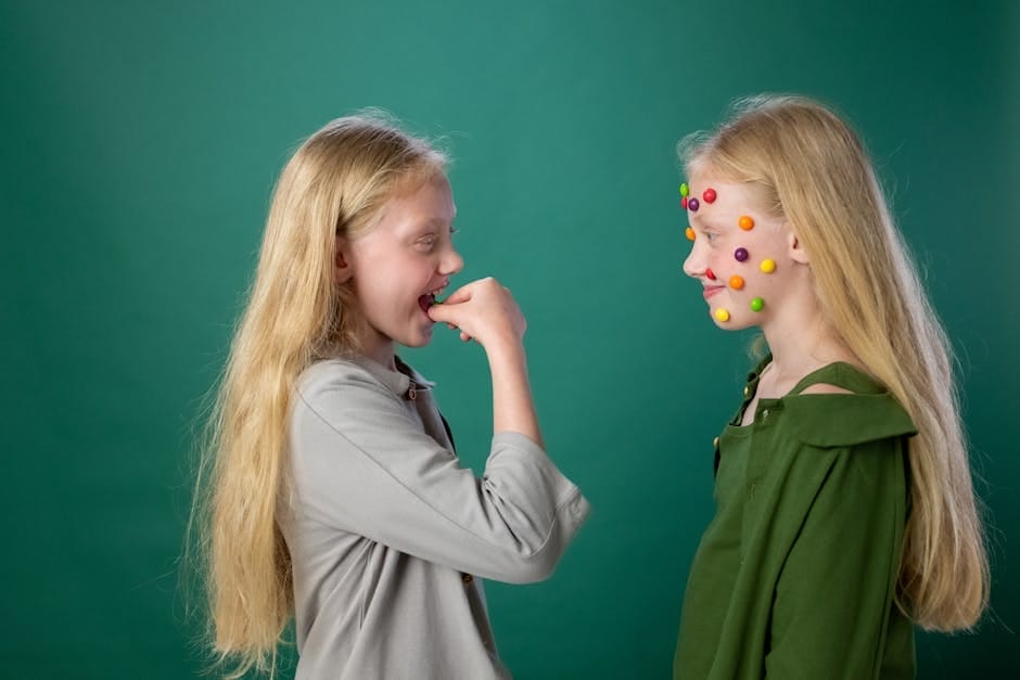 Blonde siblings having fun with colorful candy face stickers on a green background. Artistic play indoors.