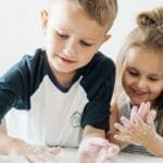 Two happy children enjoying baking, covered in flour while preparing dough at home.