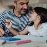 Father and daughter bonding through painting at a desk, creating art together indoors.