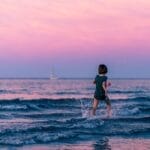 Back view of anonymous kid running along seashore and splashing water while having fun in summer at sundown