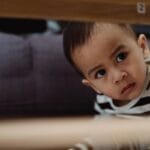 A young child in a striped shirt looks curiously from under a table, indoors.